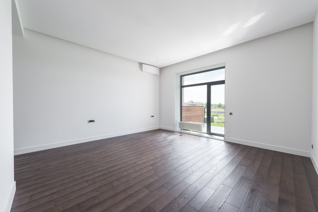 heros-img Bright and spacious modern room with a dark wooden floor and large window offering outdoor view.
